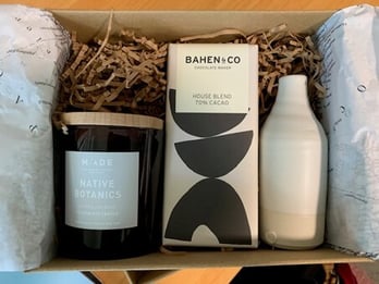 A neatly arranged gift box filled with shredded paper, featuring a scented candle, a bar of dark chocolate, and a ceramic diffuser bottle, creating a calm and thoughtful self-care set.