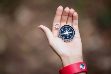 Hand holding a small black compass against a blurred outdoor background, symbolizing direction and guidance.